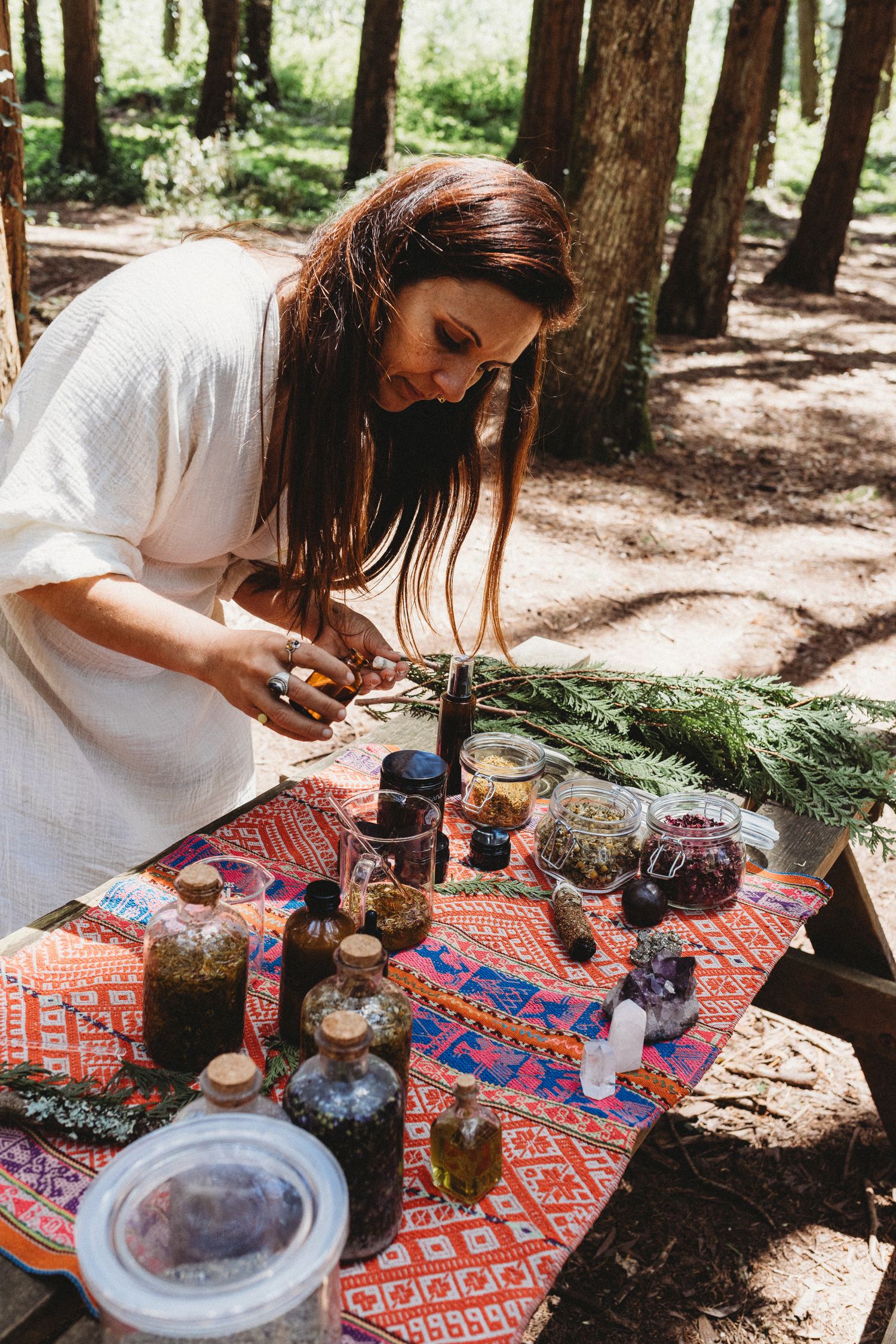 Karina at a table in the forest filling an amber bottle, surrounded by glass jars of dried chamomile and rose petals, crystals and cedar branches