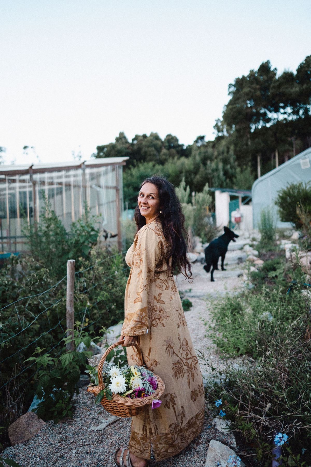 Karina in her garden at dusk, carrying a basket of freshly cut flowers, the greenhouse and her dog behind her