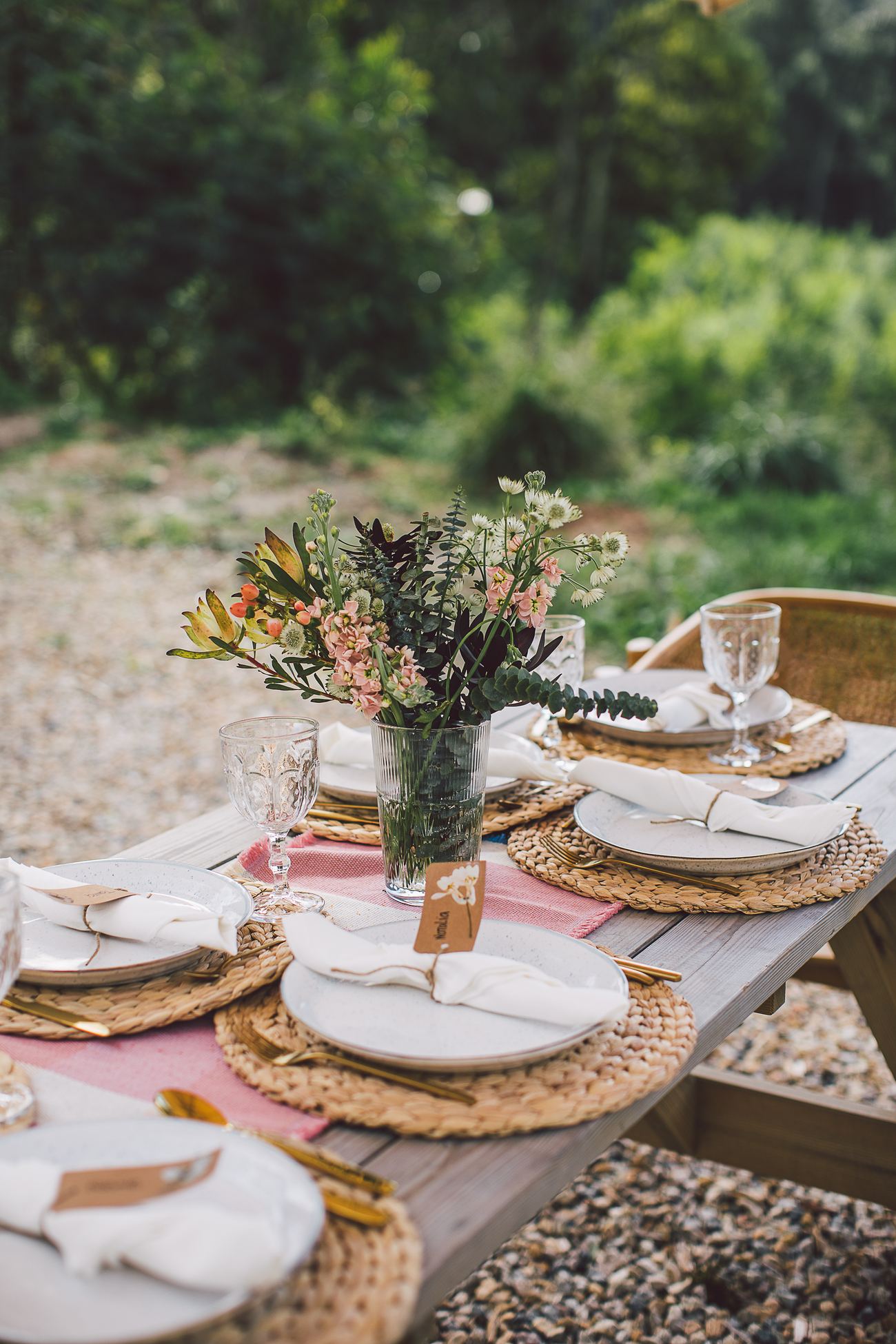 A long table set with woven placemats, ceramic plates and wildflowers, the forest behind