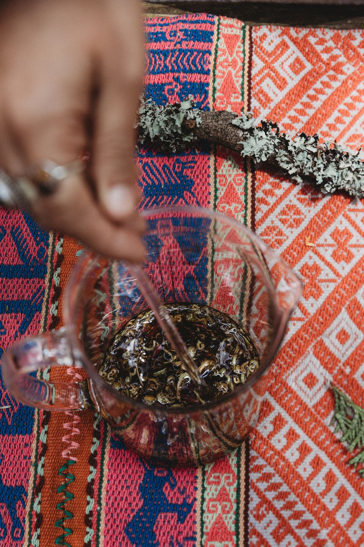 A hand stirring a dark herbal infusion in a glass teapot on a woven textile