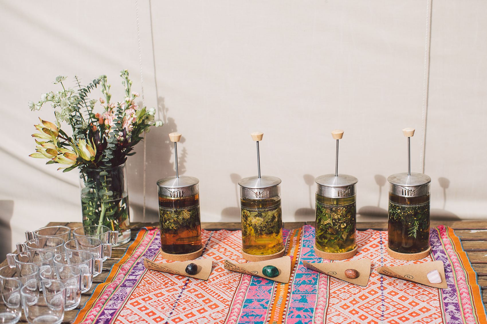 Glass tea presses filled with fresh herbs, flowers beside them